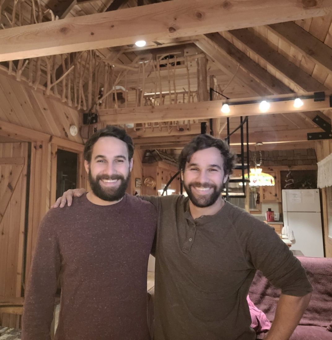 The two brothers, identical twins, standing side by side under the exposed timber frame of a cabin.